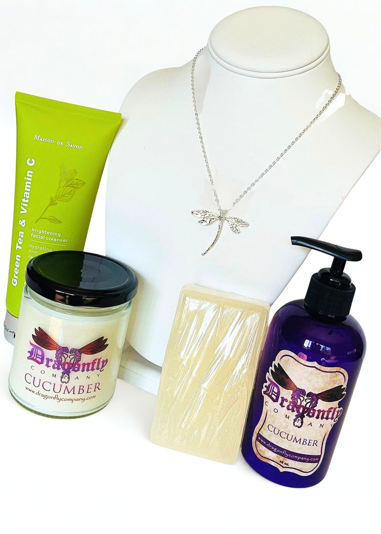 A product bundle featuring a silver dragonfly necklace, a green tube of brightening facial cleanser, a jar of Dragonfly Cucumber candle, a wrapped bar of soap, and a purple pump bottle of Dragonfly Cucumber lotion all on a white background.