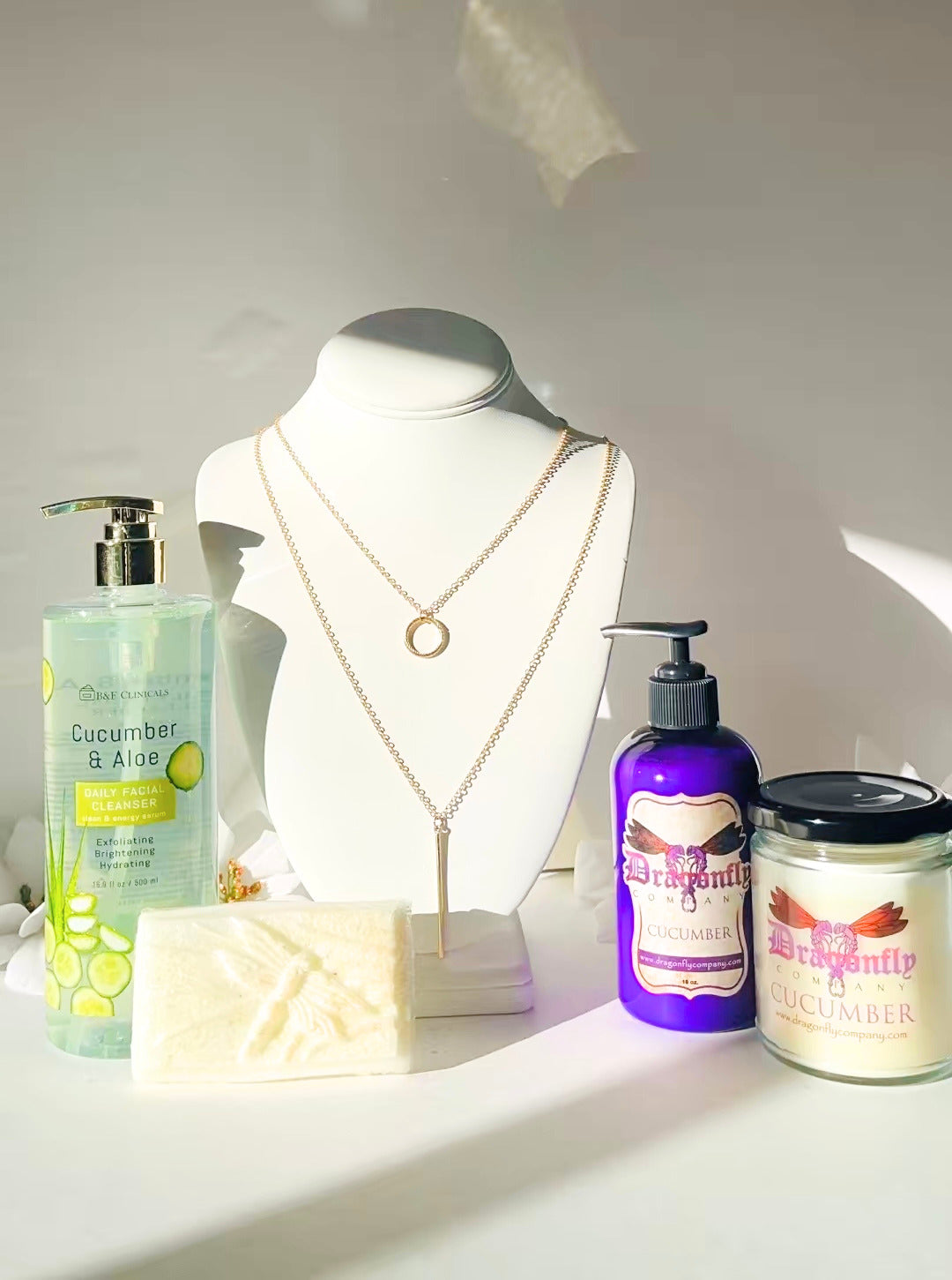 gold necklace, cucumber and aloe skincare products, a candle on a white surface.