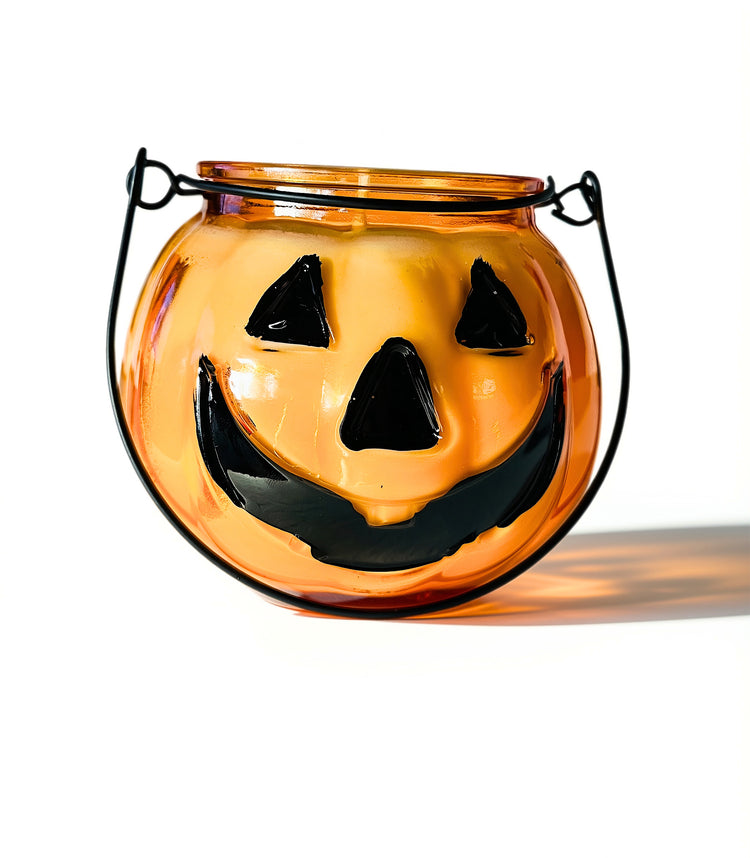 An orange glass pumpkin-candle with a black metal wire handle and a carved jack-o'-lantern face, shown on a white background with a soft shadow.