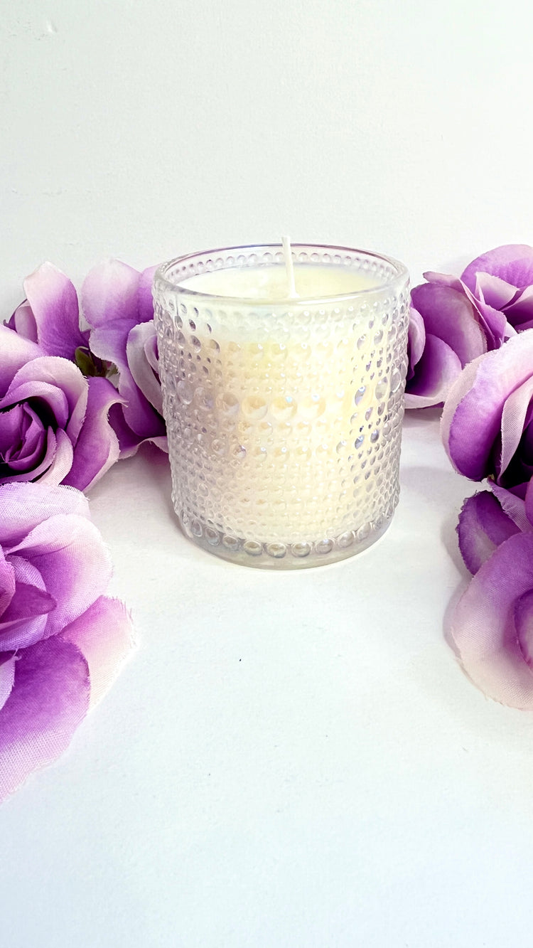 Candle in a textured glass holder surrounded by purple flowers on a white background