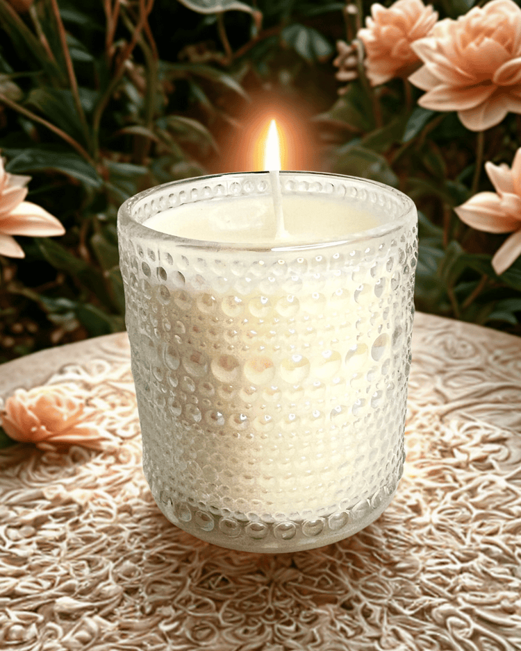 Candle in a textured glass holder on a decorative surface with flowers in the background