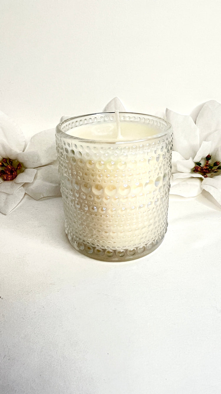 White candle in a textured glass holder with white flowers and pinecones on a light background