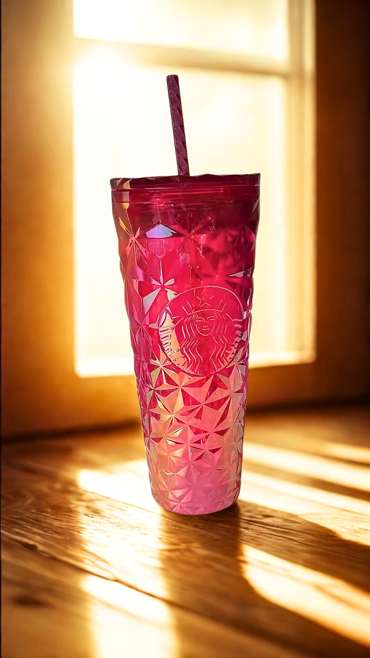 A tall, translucent pink Starbucks tumbler with a faceted geometric pattern and a dark pink straw, placed on a wooden surface in warm sunlight near a window.