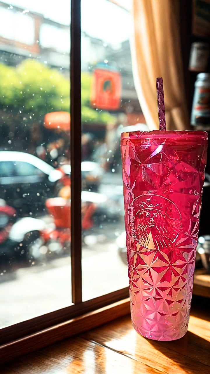 A pink Starbucks tumbler with a faceted diamond pattern and a matching pink lid and straw, sitting on a wooden table in front of a window with a blurred street scene outside.