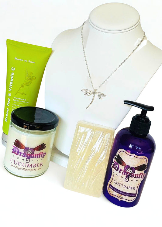 A product bundle featuring a silver dragonfly necklace, a green tube of brightening facial cleanser, a jar of Dragonfly Cucumber candle, a wrapped bar of soap, and a purple pump bottle of Dragonfly  Cucumber lotion all on a white background.