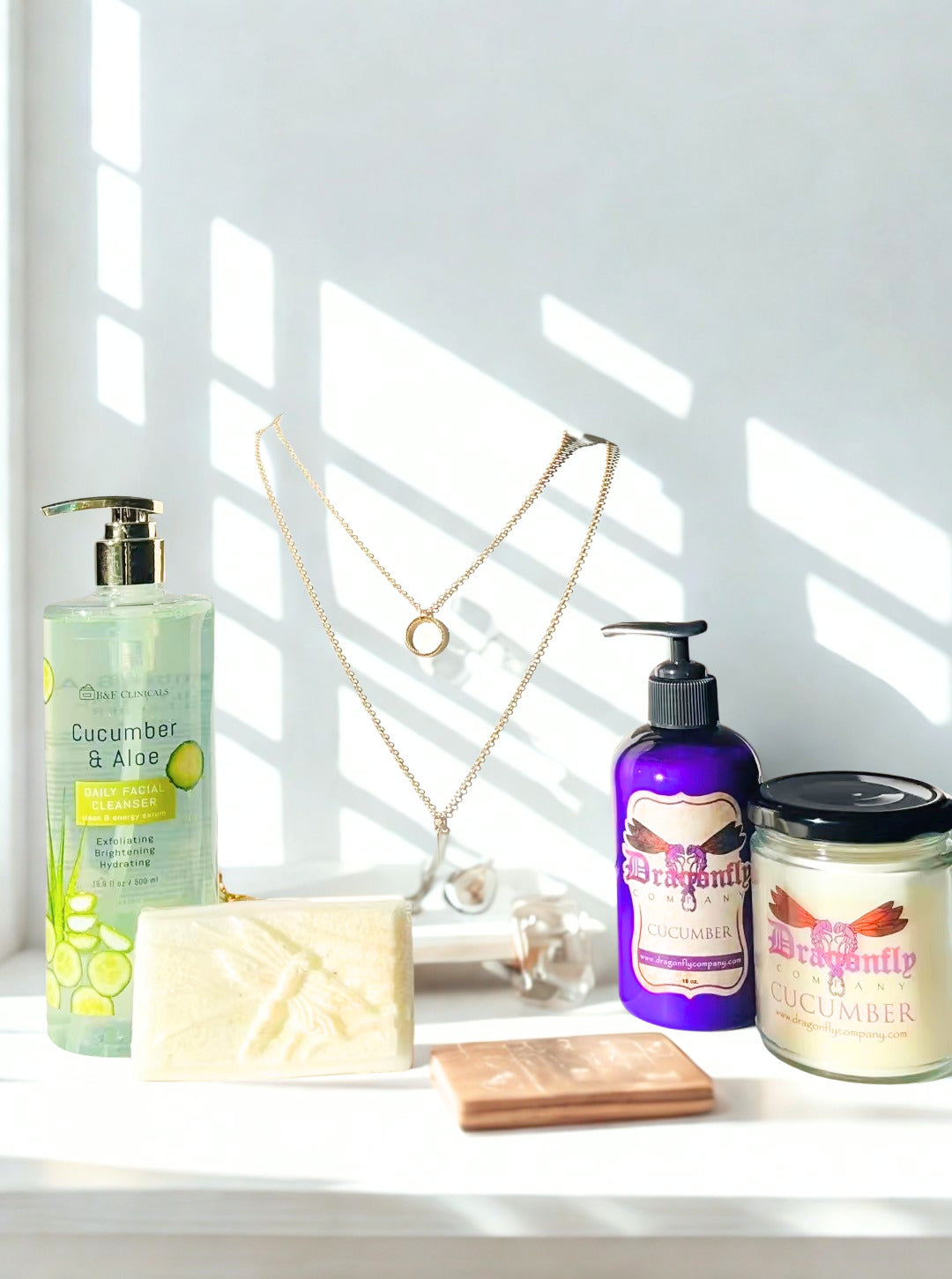 Bathroom products including a lotion, soap, and candle, facial cleanser and a necklace  on a windowsill with sunlight streaming in.
