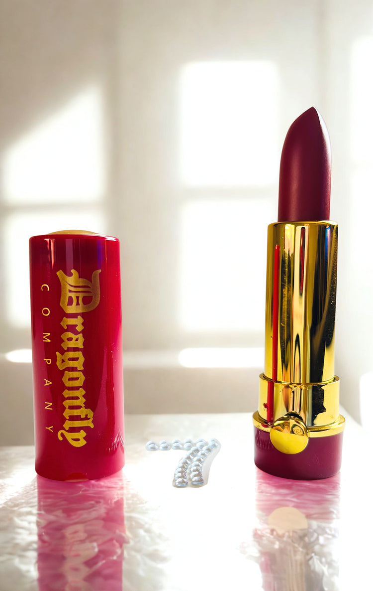 Berry  lipstick with gold cap and 'Dragonfly branding on a reflective surface.