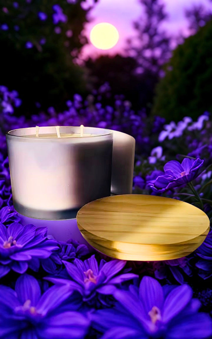 A lit  candle in a clear glass jar sits on a bed of purple flowers, with a round wooden lid placed beside it. The background is a soft-focus outdoor garden scene at dusk with purple foliage and a glowing sun.