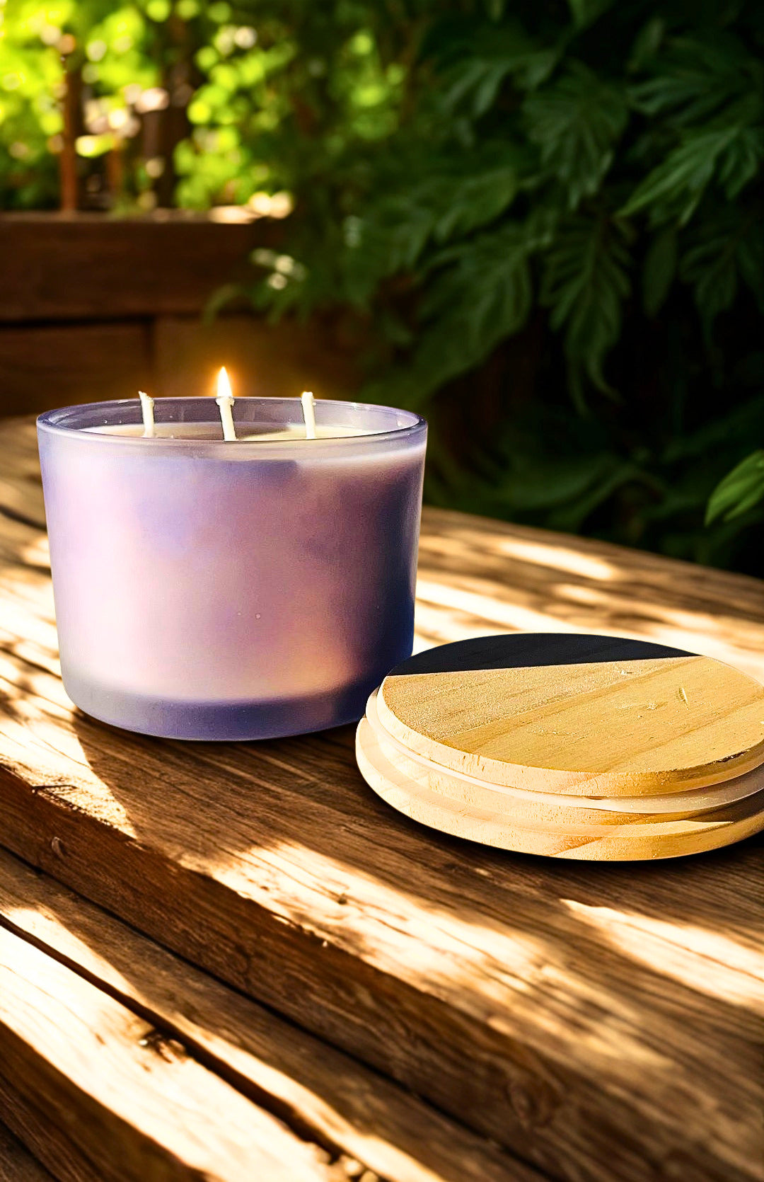 A lit lavender-colored candle in a frosted glass jar sits on a wooden table outdoors, with its round wooden lid placed beside it and blurred green foliage in the background.