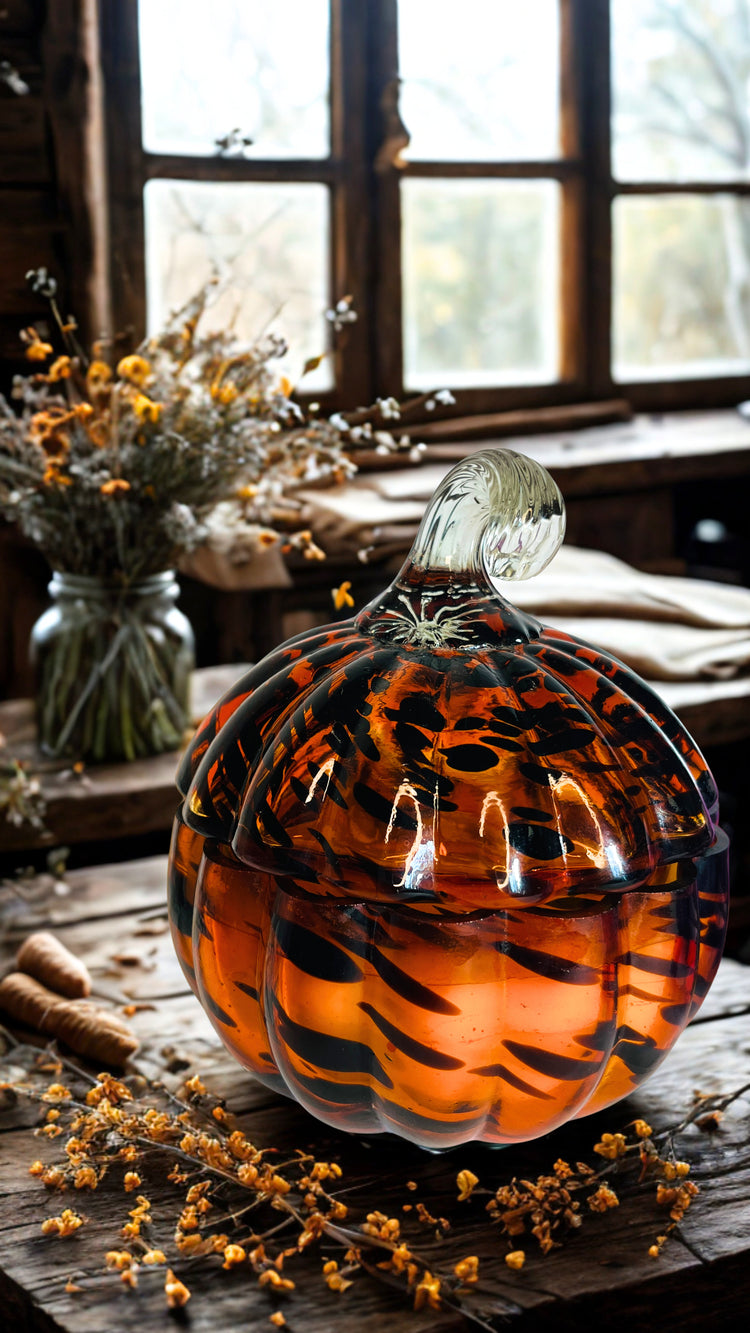 A decorative glass pumpkin candle with a ribbed pumpkin shape, filled with amber-orange  accents with dark tiger-stripe patterns, set on a rustic wooden table with dried flowers and a window in the background.