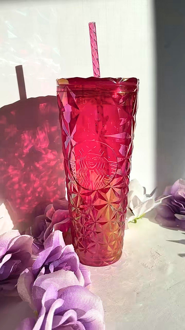 A textured, faceted pink Starbucks  tumbler with a matching pink striped straw, set against a white background with decorative purple flowers and a soft shadow.