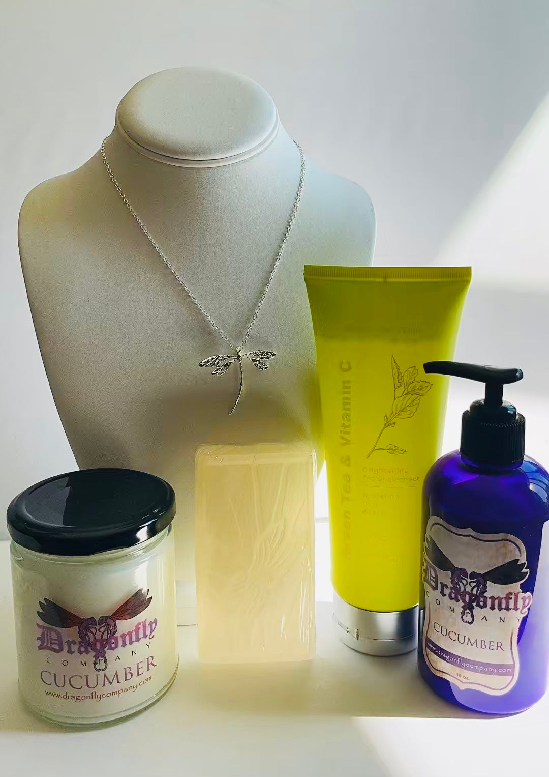 A curated collection of wellness and beauty products including a silver dragonfly necklace ,a Green Tea & Vitamin C face wash, a purple pump bottle of Cucumber lotion, a candle  and a bar of soap, arranged on a white surface.