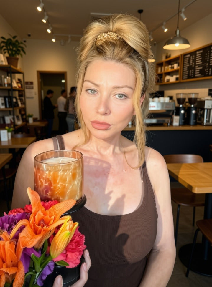 A woman in a cafe setting holds a glass jar candle with a marbled orange and amber pattern, in front of a bouquet of vibrant orange, pink, and purple flowers.