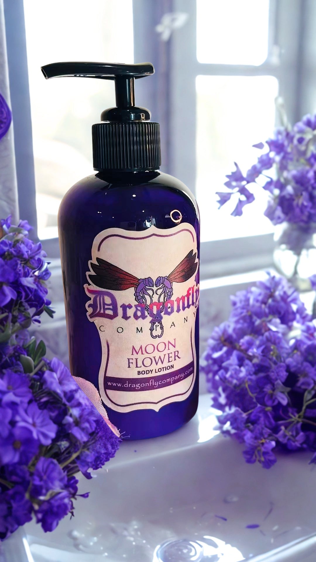 A bottle of Dragonfly Company Moon Flower Body Lotion in a deep purple bottle with a black pump, styled with purple flowers on a white windowsill.