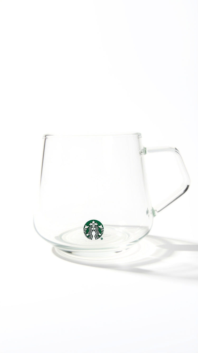 A clear glass mug with a handle and a small green Starbucks siren logo near the bottom, photographed on a clean white background with a soft shadow.