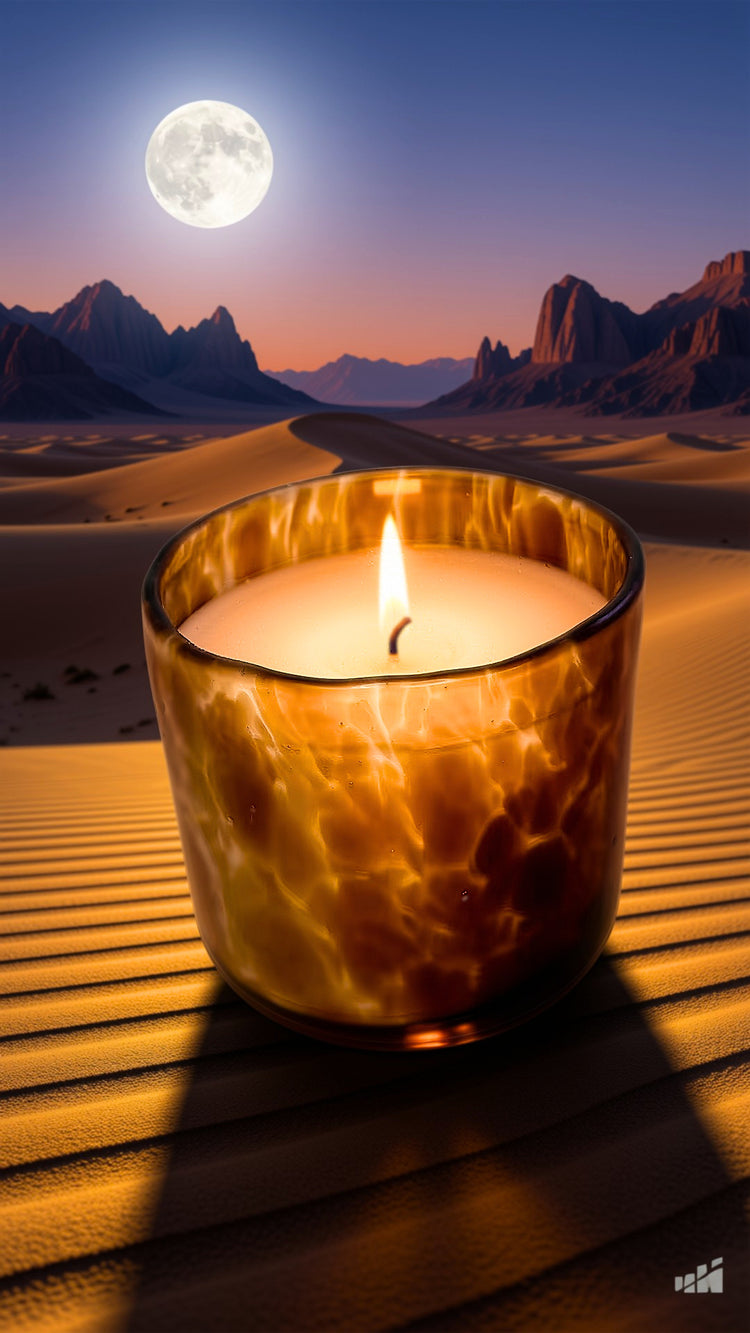 A lit candle in a textured amber glass holder sits on sand dunes with a dramatic desert landscape, mountains, and a full moon in the background.